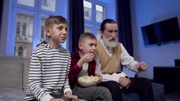Передній план захоплюючого концентрованого бородатого старшого чоловіка і двох хлопчиків-підлітків, які спостерігають за футбольним матчем на телебаченні і радіють мети — стокове відео
