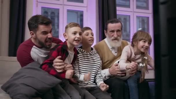 Entusiasmado pai barbudo moderno e avô sênior com três alegres netos adolescentes assistindo jogo de futebol e celebrando o objetivo — Vídeo de Stock