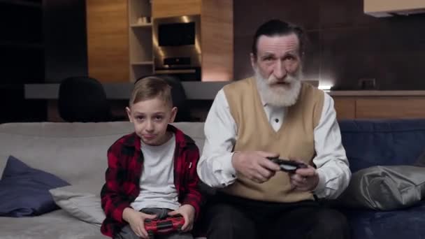 Μπροστά όψη του συγκεντρωμένου σύγχρονου γενειοφόρου ηλικιωμένου και του σοβαρού εγγονού του που παίζει μαζί βιντεοπαιχνίδι με gamepads — Αρχείο Βίντεο