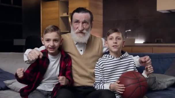 Beau-beau grand-père mature heureux avec barbe et deux petits-fils adolescents satisfaits regardant ensemble sport jeu à la télévision — Video