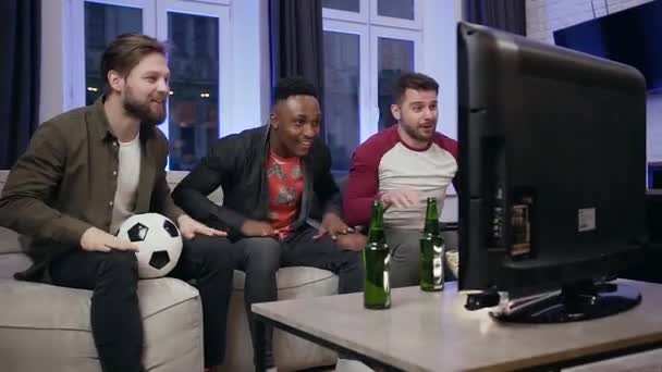 Три веселых взволнованных футбольных болельщика сидят перед телевизором и смотрят футбольный матч с криками и поднимают руки вместе — стоковое видео