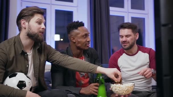 Portrait of three good-looking modern 30s multiethnic guys which eating popcorn during watching sport game on tv and talking about actions of players — ストック動画