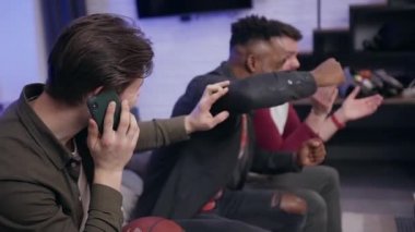 Attractive agitated diverse two young guys emotionally watching sport game and interfering their third bearded mate to talk on phone