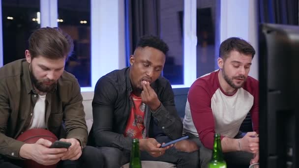 Front view of good-looking modern sad young diverse men which using their phones during watching sport game on tv — Stok video