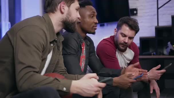 Side view of good-looking serious modern young diverse guys which sitting on sofa and using their phones while watching tv programm — Stock videók