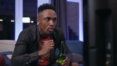 Attractive concentrated black-skinned guy eating chipps during watching football game and making active movements like a player