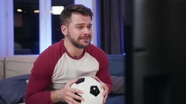 Good-looking excited 30-aged bearded man holding football ball in his hands during watching football match on tv and disappointing putting his head on ball as his favourite team lose a chance