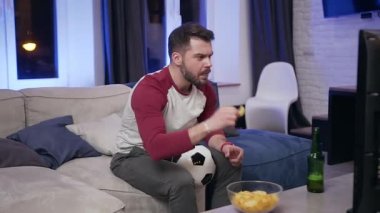 Handsome concentrated bearded young man eating chipps during reviewing the football match on tv at home