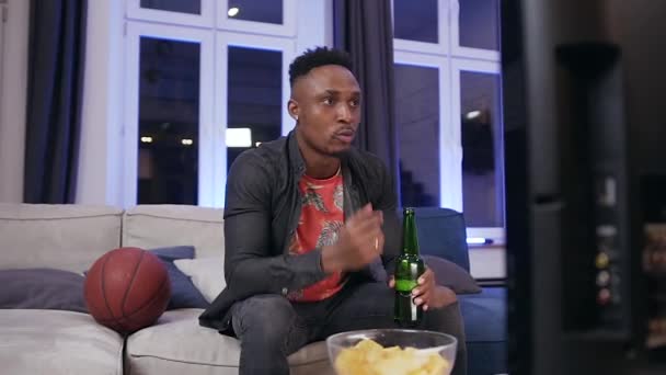 Good-looking concentrated african american eating chipps,drinking beer while emotionally watching football match on tv — Stok video