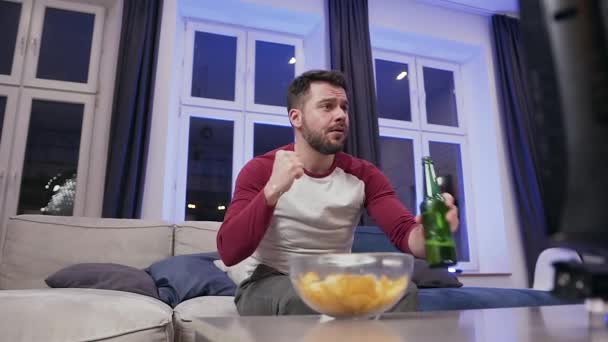 Front view of attractive excited 30-aged bearded man which encouraging his favourite team on tv and enjoying beer together with chipps — Stock video