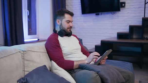 Handsome smiling satisfied 30-aged guy sitting on the couch and browsing apps on i-pad — Αρχείο Βίντεο
