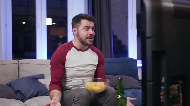 Guapo joven barbudo emocionado viendo el deporte juego en la televisión en la habitación en casa y emocionalmente comentando la obra periódicamente comer chipps — Vídeo de stock