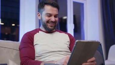Front view of smiling satisfied young bearded guy which sitting on the couch and using tablet pc for review funny videos