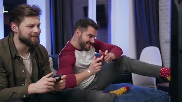 Веселые смешные 30-х бородатые парни сидят на диване и играют в видеоигры по телевизору с помощью геймпадов — стоковое видео