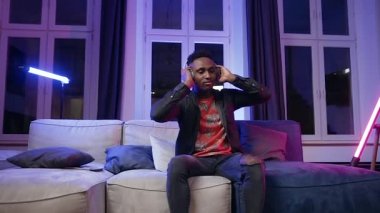 Front view of handsome trendy 30-aged african american which sitting on the violet light room background and enjoying modern music in headphones