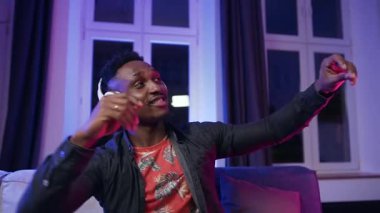 Good-looking satisfied trendy 30-aged dark-skinned guy in headphones dancing with hands under cool disco music ,sitting on the sofa in the room with violet light