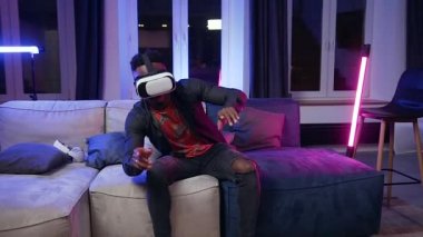 Attractive smiling young black-skinned man in virtual reality headset sitting on the couch in the contemporary room in the evening time
