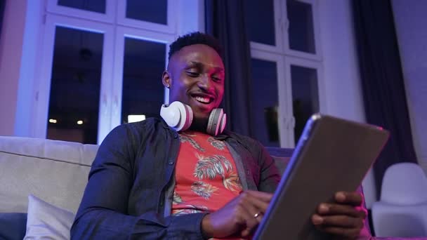 Привлекательный современный позитивный африканский американец, сидящий на диване и смотрящий смешные картинки на своем i-pad — стоковое видео