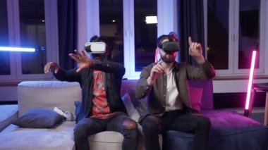 Attractive positive modern multiracial male friends sitting on the couch in the room and making hands activities in air as on virtual screen using special 3d glasses