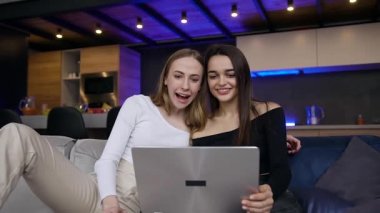 Çağdaş odadaki rahat koltukta ortak arkadaşlarla bilgisayar başında video görüşmesi yapan neşeli, çekici ve pozitif genç bayanların ön görüntüsü.