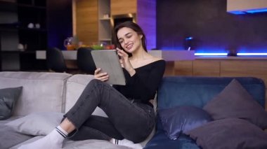 Çekici, modern, siyah bluzlu, kot pantolonlu esmer kadın. Kanepede oturuyor ve tablet bilgisayar kullanıyor.