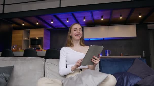 Вид спереди улыбающейся симпатичной довольной девушки в белой одежде, которая сидит на диване и делает удивленное лицо во время работы над i-pad — стоковое видео
