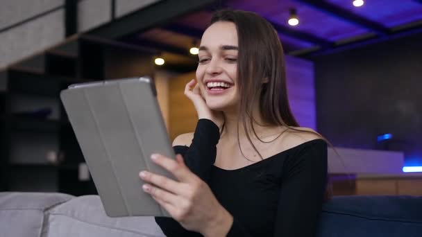 Boa índole positiva menina alegre emocional feliz sorrindo durante a revisão de suas fotos no tablet pc, sentado no sofá em casa — Vídeo de Stock