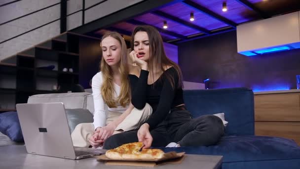 Attraktive positive oppspilte 25-ere som sitter på sofaen og spiser pizza under videolesing på datamaskinen – stockvideo