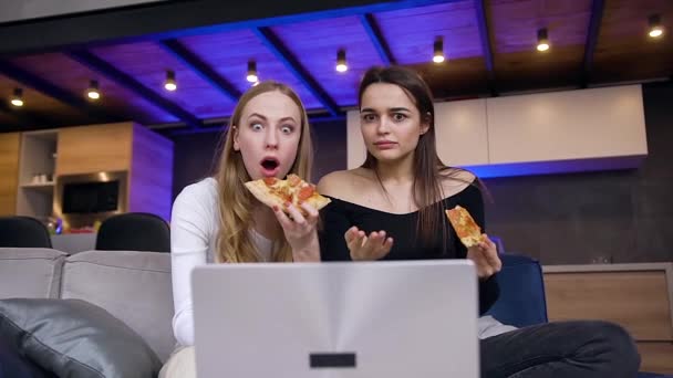 Közel a szerethető vidám fiatal barna és szőke, amely ellazult a kanapén, eszik pizza közben vicces videókat a laptopon — Stock videók