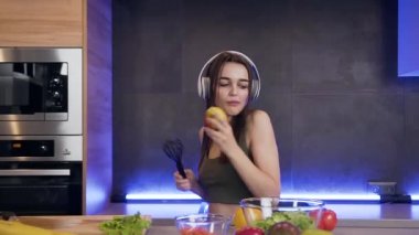 Güzel, enerjik, ince kulaklıklı genç bir kadın mutfakta dans ederken taze elma yerken müzik dinliyor.