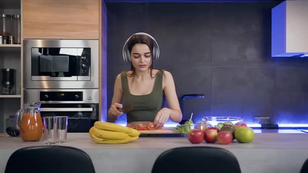 Jovem alegre e alegre que está perto da mesa da cozinha e cortando tomate vermelho para salada de legumes enquanto ouve música em fones de ouvido — Vídeo de Stock