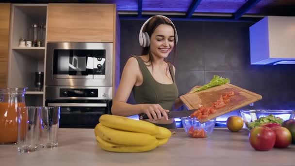 헤드폰을 끼고 현대적 인 미소짓기 좋아 보이는 갈색 수염을 닫고 야채 샐러드를 준비하는 동안 그릇에 얇게 썬 토마토를 넣는다. — 비디오