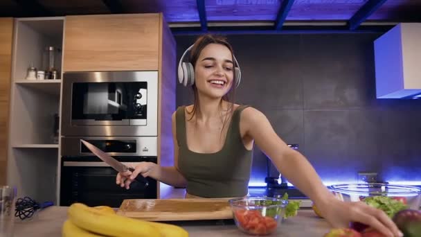 Közel a csinos mosolygós boldog fiatal nő, amely eszik almát, miközben zenét hallgat a fülhallgató a konyhában — Stock videók
