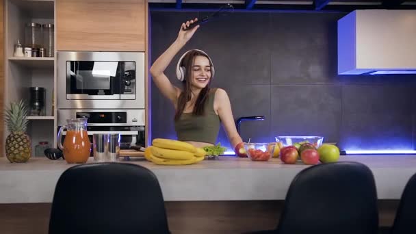 Vista frontal de la risa divertida joven bonita que baila bajo la música en sus auriculares en el fondo de la cocina bellamente diseñado — Vídeo de stock