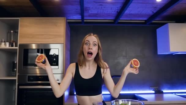 Позитивная игривая улыбающаяся молодая женщина держит две половинки грейпфрута в руках и делает с ними очки на глазах — стоковое видео