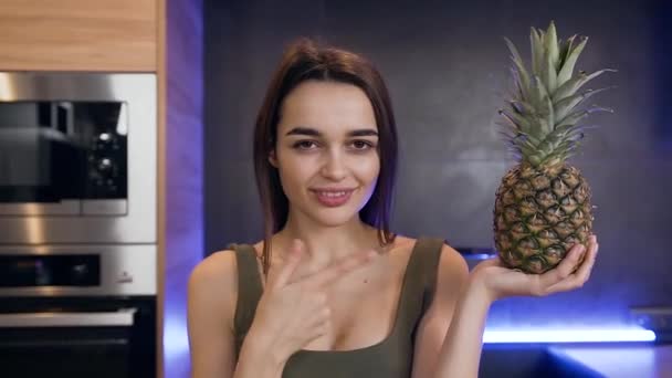 Vrij tevreden moderne jonge brunette houden in haar hand ananas en het maken van teken "zijn goede keuze" op de keuken achtergrond — Stockvideo