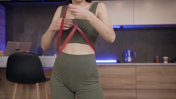 Onherkenbare slanke jonge vrouw in fitnesswear meten van haar lichaamsmaten met tape staan bij de tafel in de hedendaagse keuken — Stockvideo