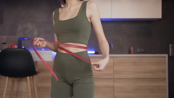 Приваблива щаслива струнка молода жінка вимірює розмір талії стрічкою і танцює від задоволення на сучасній кухні — стокове відео