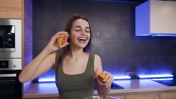 Vista frontal de sonriente morena joven delgada satisfecha que sostiene dos mitades de pomelo en el fondo de la cocina — Vídeo de stock