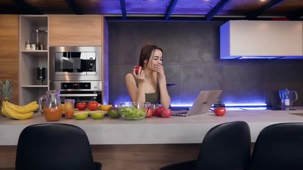 Высокопоставленная позитивная молодая женщина ест свежее яблоко и пользуется ноутбуком на хорошо оборудованной кухне — стоковое видео