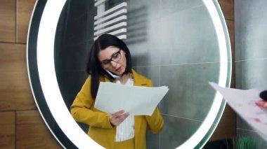 Çekici, enerjik, genç bir iş kadını aynanın karşısında durmuş, telefonla konuşurken dokümanları izliyor ve yüz estetiği kullanıyor.