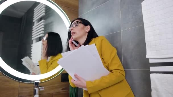 Красивая профессиональная стильная молодая брюнетка в офисной одежде разговаривает по телефону у зеркала ванной комнаты во время чтения документов — стоковое видео