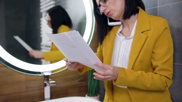 Goed uitziende elegante jonge vrouwelijke manager zit in de buurt van de grote spiegel in de badkamer met documenten en praten aan de telefoon — Stockvideo