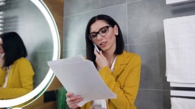 Kendine güvenen, gülümseyen, profesyonel, siyah saçlı, ofis yöneticisi, bayanlar tuvaletindeki yuvarlak aynanın yanında yaptığı telefon görüşmeleri sırasında mali raporları okuyor.