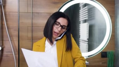 Ofis kıyafetleri içinde şık modern moda genç iş kadını banyoda aynanın yanında duruyor ve finansal raporları okurken telefonla konuşuyor.