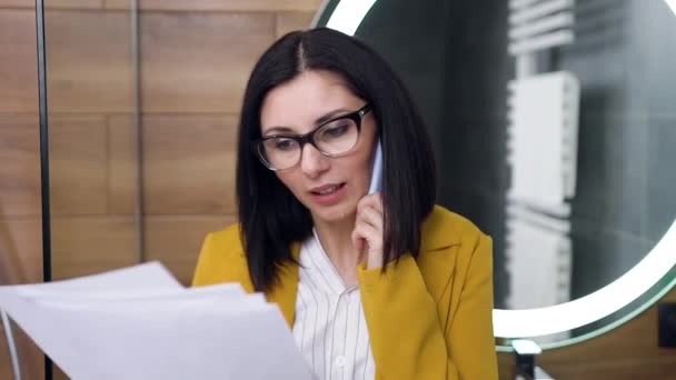 Primo piano di attraente elegante giovane donna d'affari che in piedi vicino al grande specchio ed emotivamente parlando con l'interlocutore su smartphone, la lettura di documenti finanziari — Video Stock