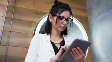 Beyaz elbiseli, i- pad 'inden finansal raporlar okuyan, evdeki büyük aynanın yanında duran, göz kamaştırıcı genç iş kadınının ön görüntüsü.