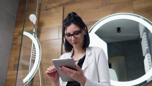 Apelando elegante mujer de negocios de moda de 30 años en traje blanco de pie cerca del espejo en el baño y leyendo noticias en su i-pad y haciendo cara sorprendida — Vídeos de Stock