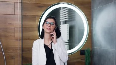 Gülümsemenin ön görüntüsü 30 yaşında beyaz takım elbiseli çekici bir iş kadınının ev banyosunda mobil sohbetleri var.