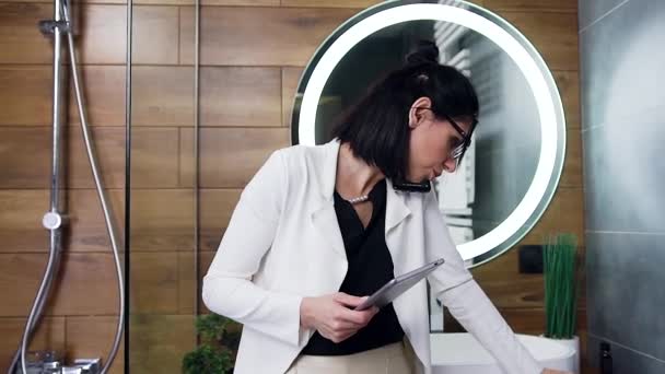 Серьёзно элегантная красивая молодая женщина с черными волосами в белом костюме разговаривает по телефону и читает финансовый отчет в своей ванной комнате — стоковое видео